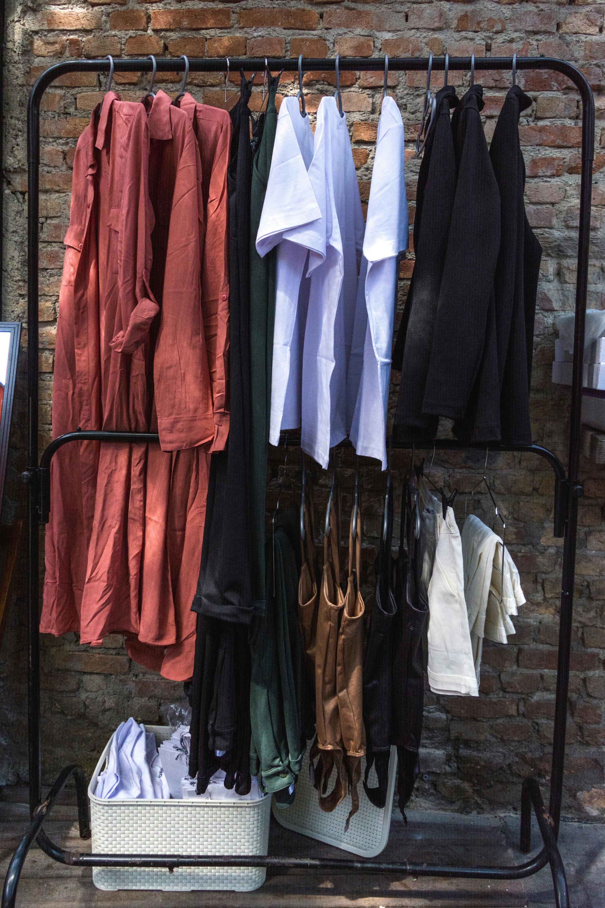 Clothes rack with various garments: dresses, shirts, tops in pink, white, black, green, and brown against a brick wall.