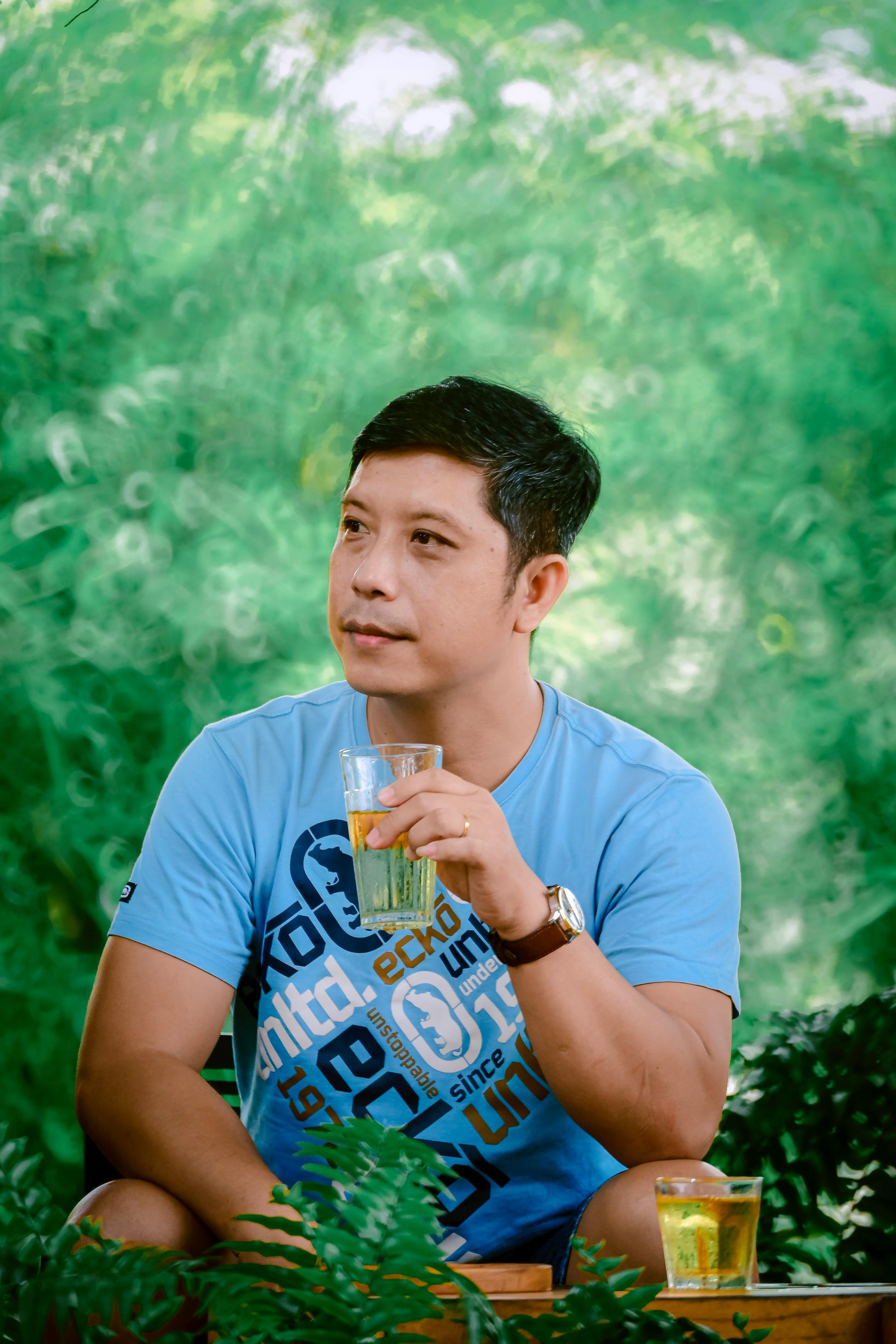 Man in blue shirt holding drink, wearing watch, outdoors with green background.