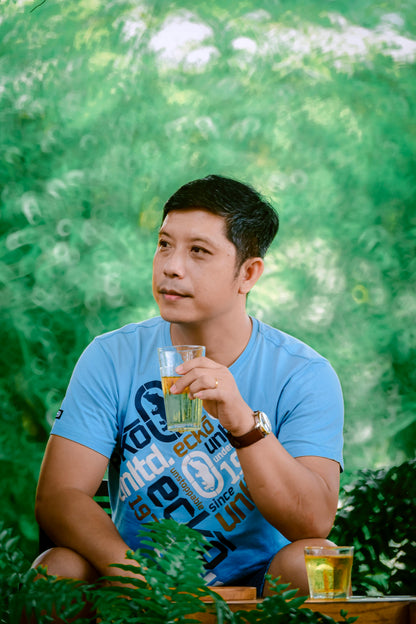 Man in blue shirt holding drink, wearing watch, outdoors with green background.